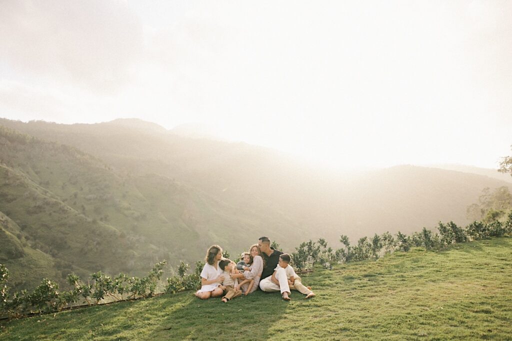 A family of 6 sit together and cuddle on a cliff looking out over a mountain of Oahu during their family session