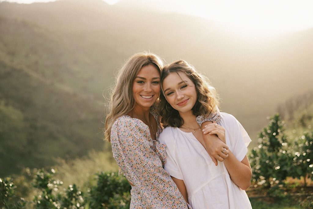 A mother and her daughter pose and smile at the camera during their family session on Oahu, behind them the sun is rising over a mountain