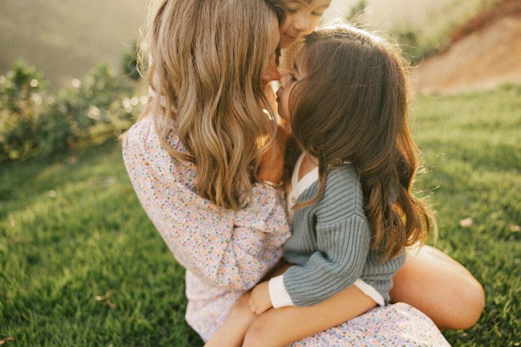 A mother touches her nose to her youngest daughter's nose while her youngest son hugs her from behind, the sun is rising behind them