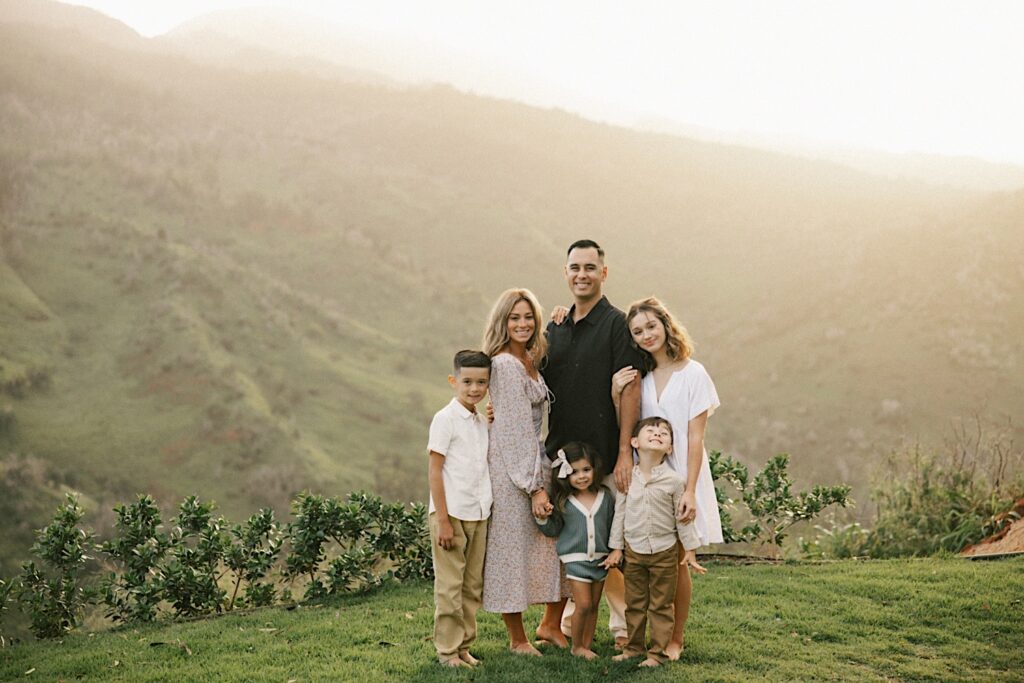 During a family session, a family of 6 stand and pose together smiling at the camera while the mountains of Oahu are covered in fog behind them