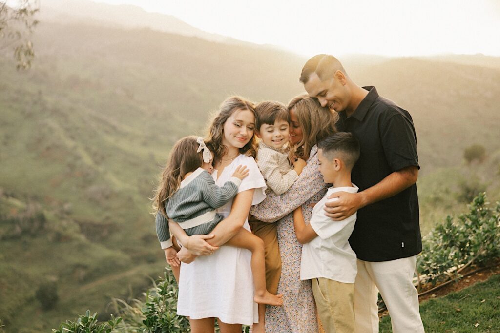 During a family session, a family of 6 stand and embrace while smiling at one another while the mountains of Oahu are covered in fog behind them
