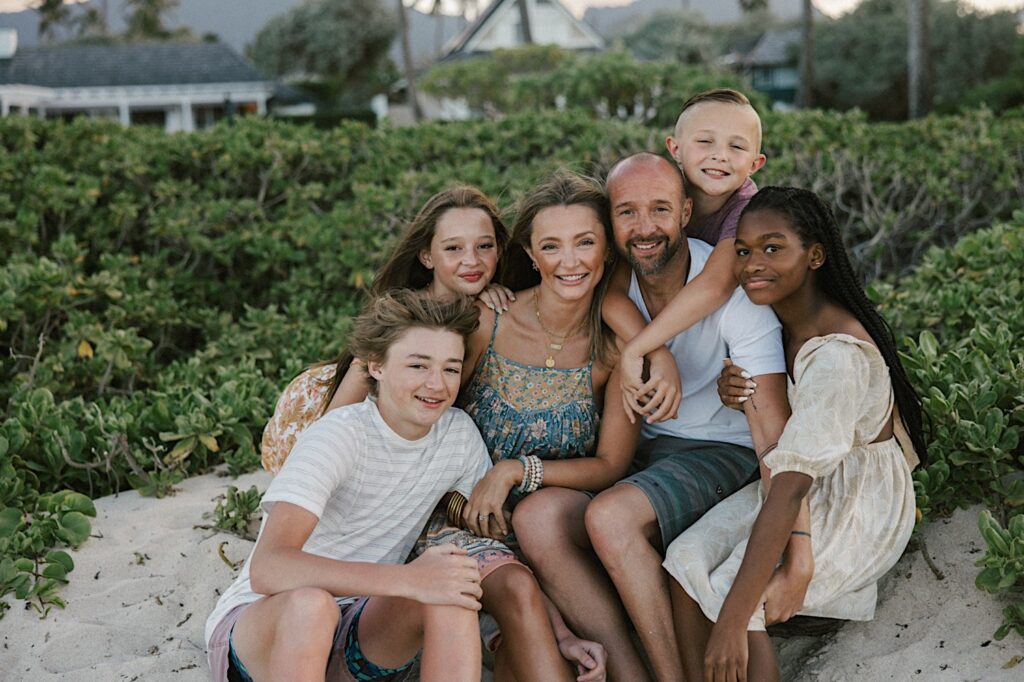 A family of six sit together on a beach of Hawaii and smile at the camera during their session, greenery and buildings are in the background behind them