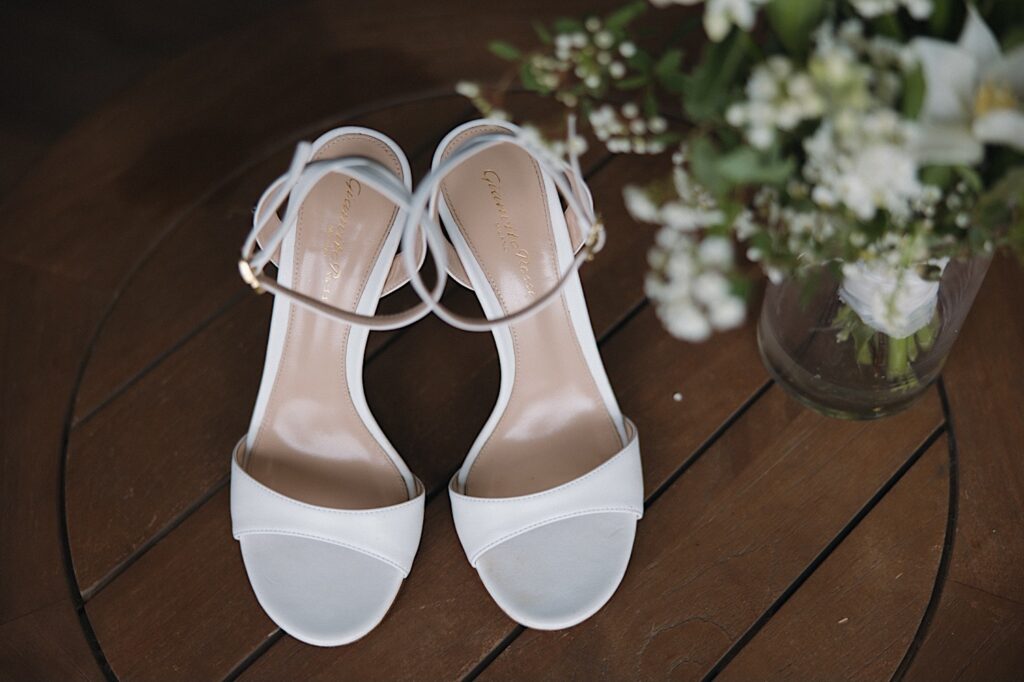 Detail photo of a bride's wedding shoes on a table next to a vase with flowers in it