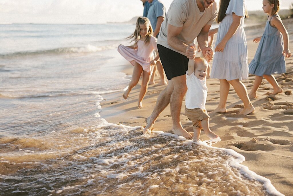 A child's hands are held by his father as they walk along the beach with the rest of their family behind them during their family session in Hawaii
