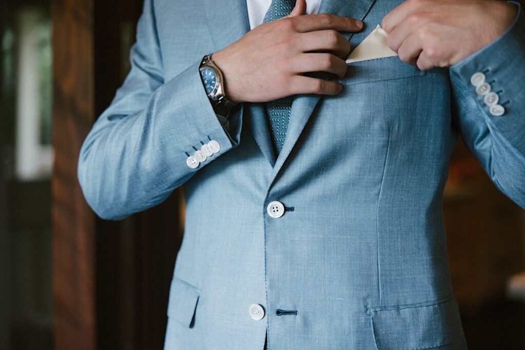 Close up photo of a groom wearing a suit and adjusting his pocket handkerchief 