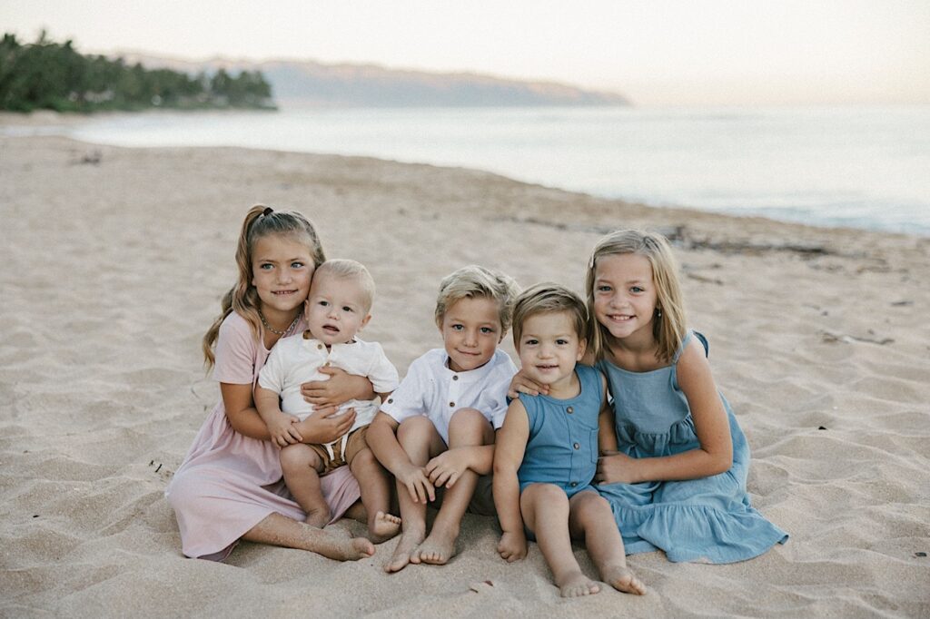 Five children sit together on a beach in Hawaii and smile at the camera with the ocean behind them during their family session