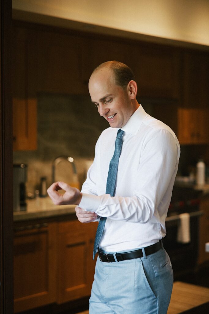 A groom smiles as he stands in a room while looking down and adjusting the cuff of his button up shirt
