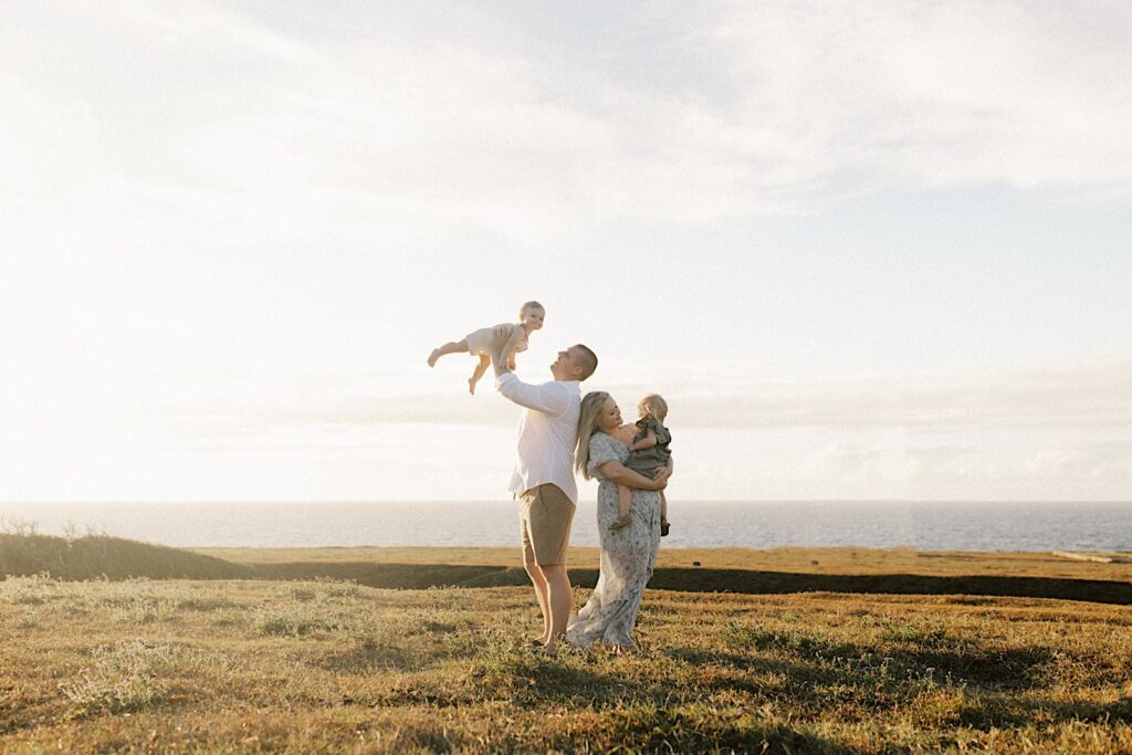 A family of four stand in a field in Hawaii looking over the ocean during their family session at sunset, the mother is holding one child while the father tosses the other in the air