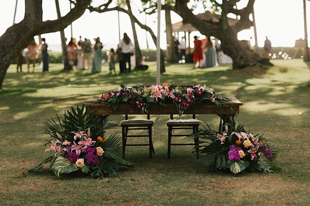 A table set up with floral decorations on it, in the background are guests of an outdoor wedding reception in Hawaii