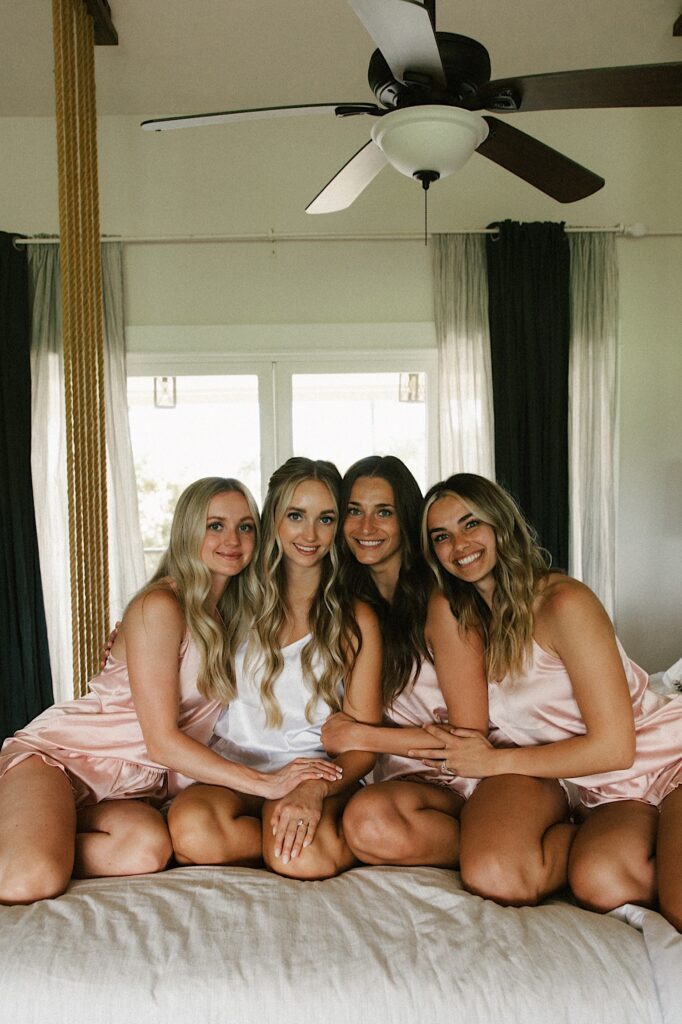 A bride and her three bridesmaids sit on a bed with one another and smile at the camera before getting ready for a wedding