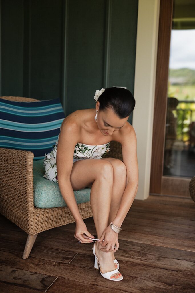 A bride sitting in a wicker chair puts on her shoes before her wedding day
