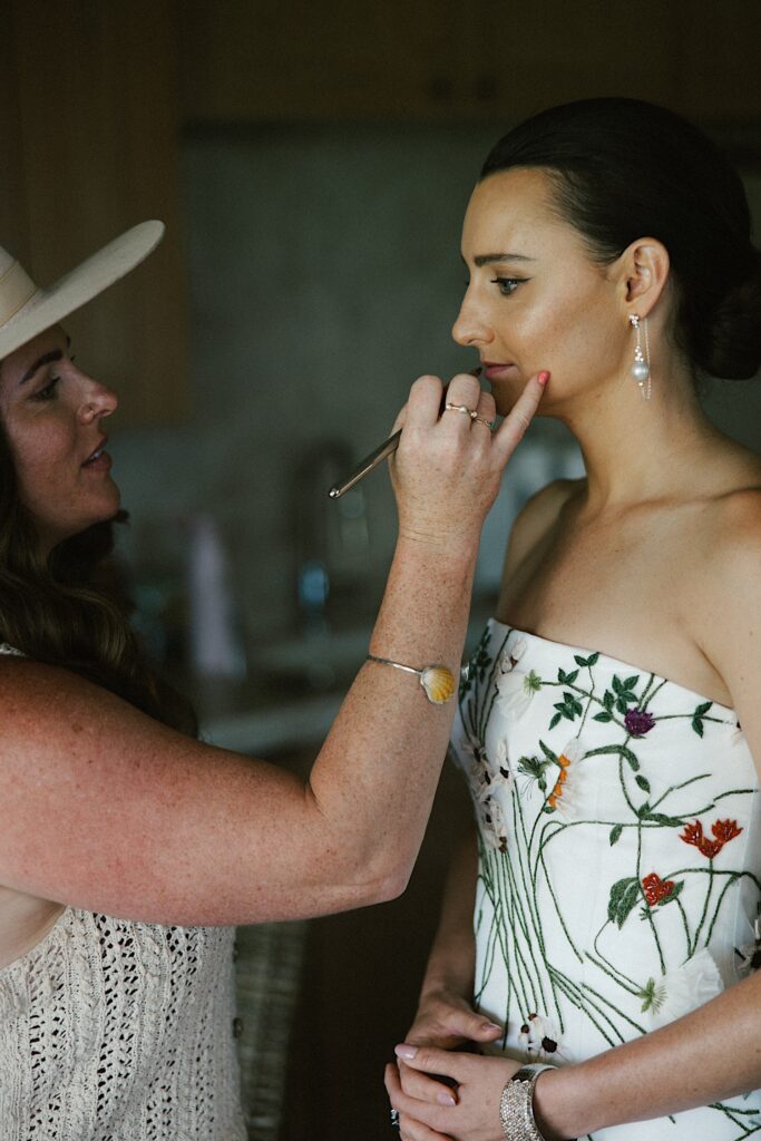 A bride stands as a woman in front of her puts her lipstick on