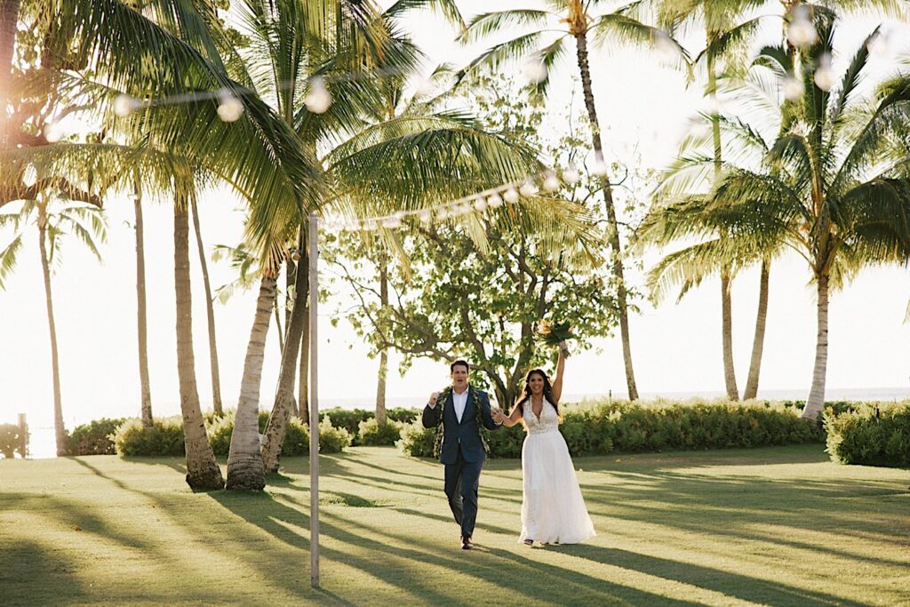 A bride and groom walk together across a field with palm trees and the ocean behind them, they are entering their wedding ceremony space at their venue Lanikuhonua on Oahu