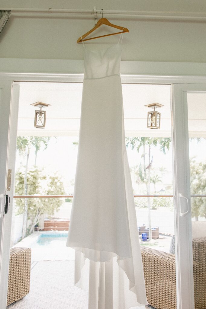 A wedding dress on a hanger hands from the ceiling in front of a glass door that leads outside