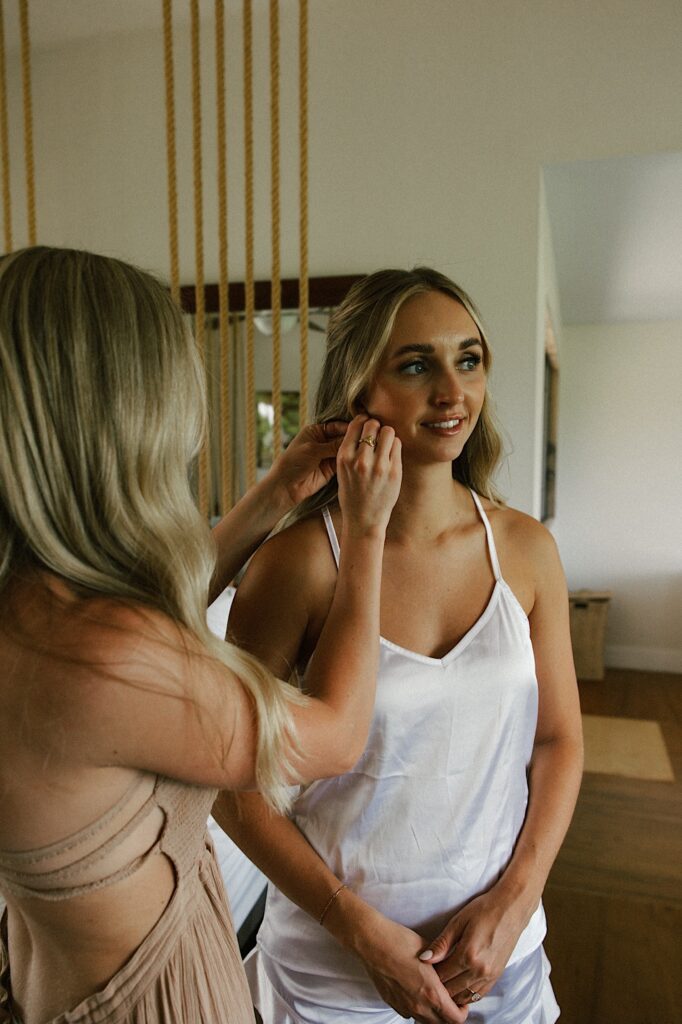 A bride stands and smiles as one of her bridesmaids helps put an earring on her