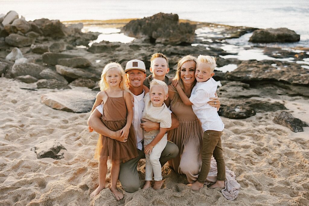 A family of six pose together on a beach of Hawaii with the ocean behind them and smile at the camera during their family session