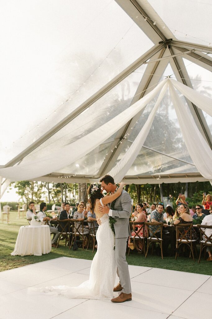 A bride and groom share their first dance underneath a large tent as guests of the wedding sit and watch