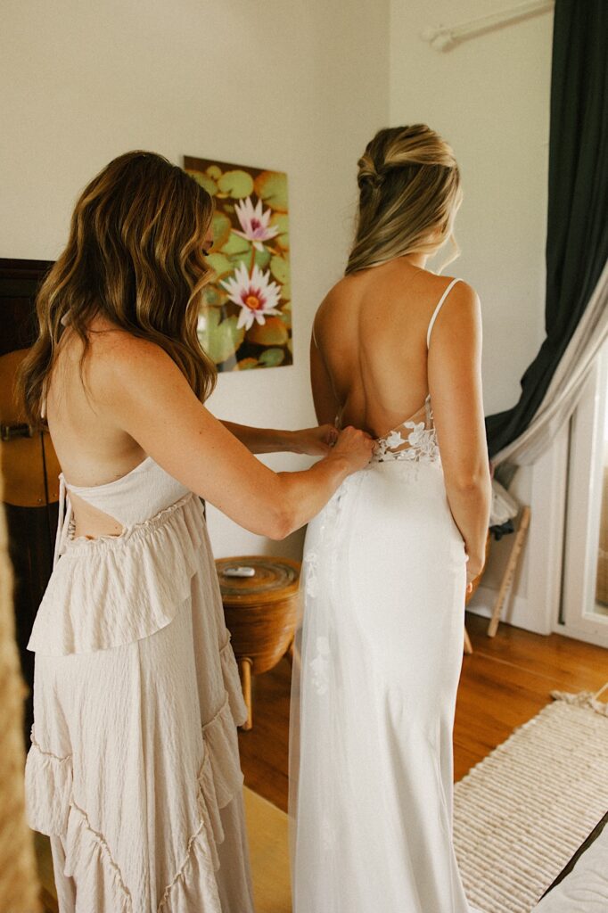 A bridesmaid stands behind the bride and helps zip up the back of her wedding dress as they get ready for a wedding