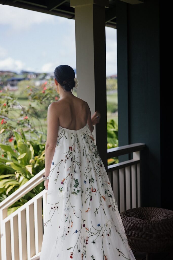A bride facing away from the camera stands on a porch and looks out over the lush greenery of Hawaii