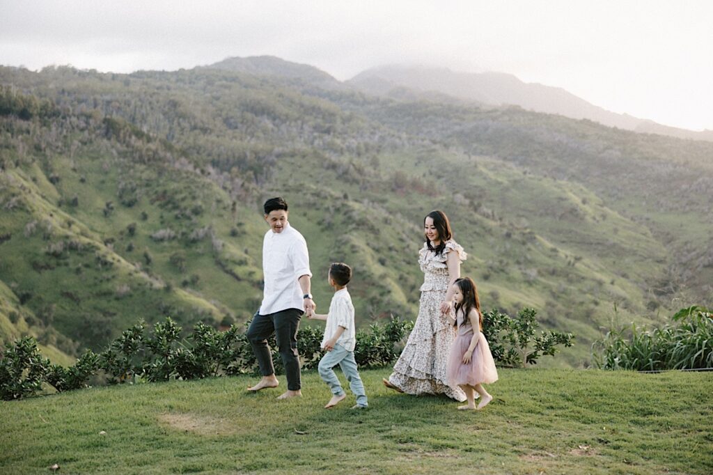 A family of four walk along a cliff in Hawaii looking over a green mountain during their family session