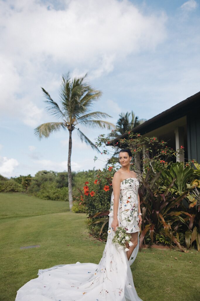 A bride stands in front of a house with lush greenery outside and looks out over her shoulder