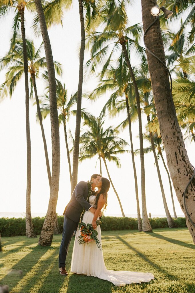 A groom kisses a bride on the cheek, they're surrounded by palm trees and the sun is setting behind them