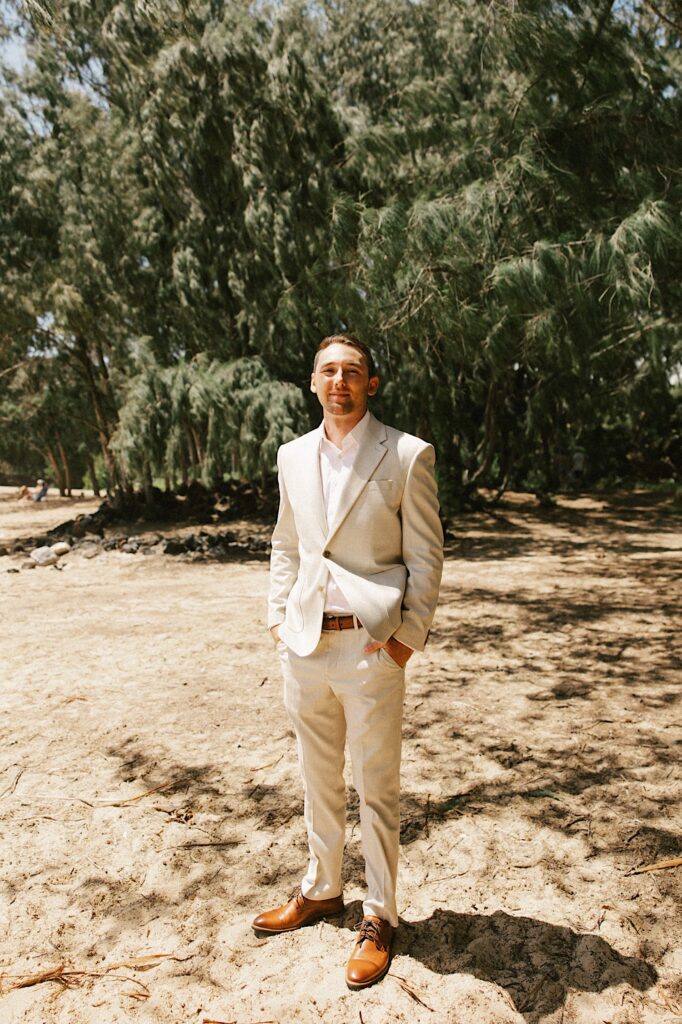 A groom stands on a beach dressed for a wedding and smiles at the camera with his hands in his pockets