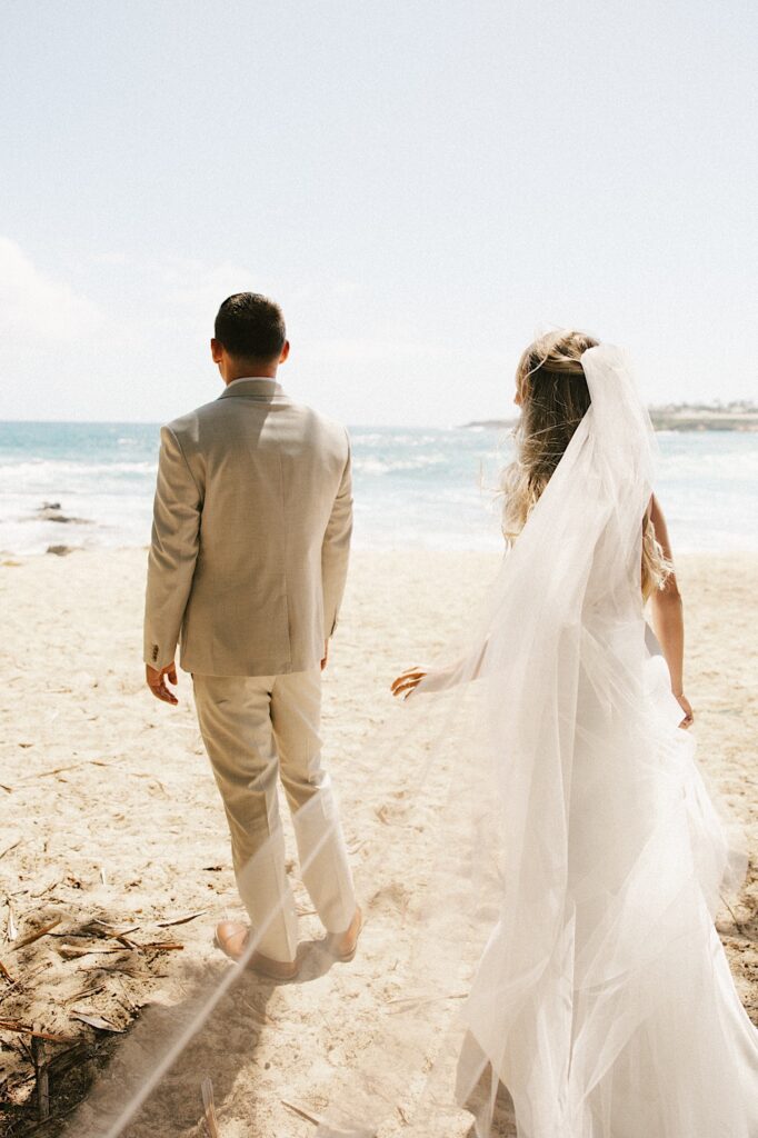 A groom stands facing away from the camera looking out over the ocean while on a beach, behind him the bride in her wedding dress is sneaking up on him to tap his shoulder before their first look