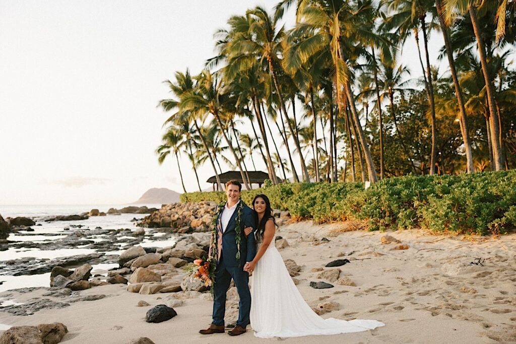 A bride and groom stand on the beach and pose in front of palm trees and the ocean near their wedding venue Lanikuhonua on Oahu