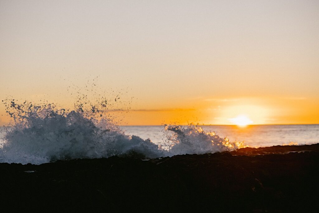 The sun sets on the ocean as a wave crashes on the shore