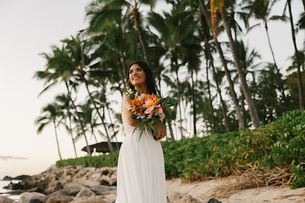 A bride smiles and looks off in the distance while on a beach in front of palm trees near her wedding venue Lanikuhonua on Oahu