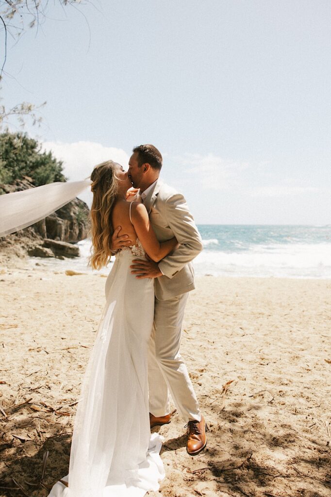 A bride and groom on a beach in their wedding attire kiss one another and embrace with the ocean behind them