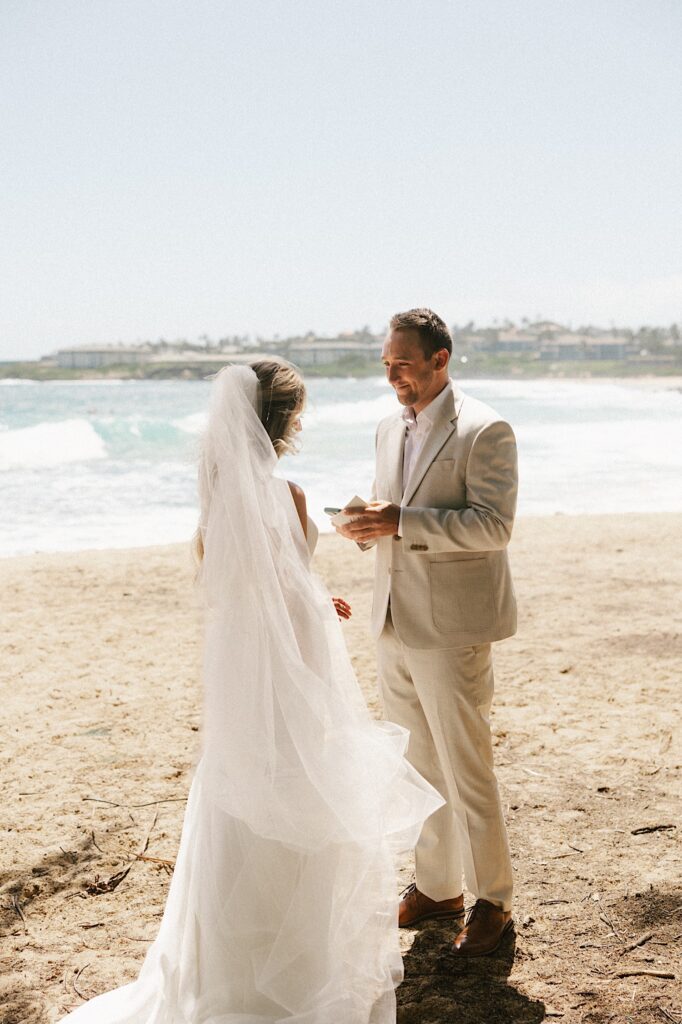 A bride and groom stand on a beach facing one another in their wedding attire with the ocean behind them, the groom is holding a book with his vows in it