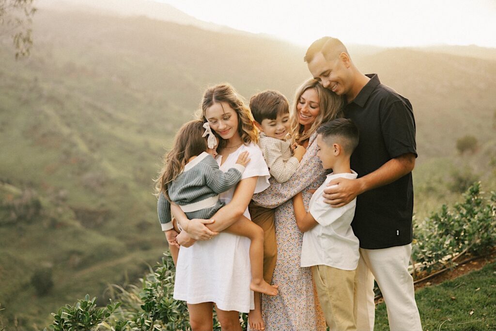A family of 6 all stand with one another and pose while standing in front of the mountains of Hawaii during their family session, the mother is holding the youngest son while the oldest daughter is holding the youngest daughter 