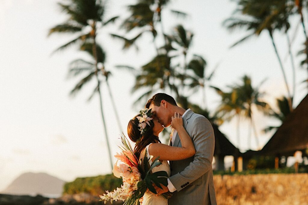 A bride and groom kiss on the beach with palm trees behind them near their wedding venue Lanikuhonua on Oahu