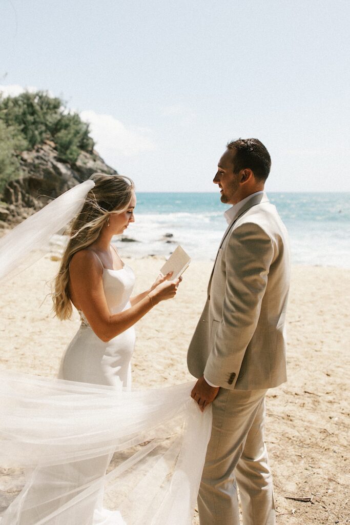 A bride reads her vows to the groom standing in front of her as he holds her veil while the two stand on a beach with the ocean behind them