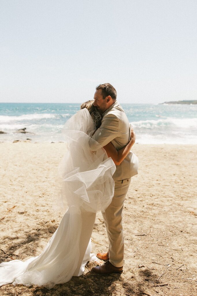 A bride and groom on a beach with the ocean behind them hug with the ocean behind them