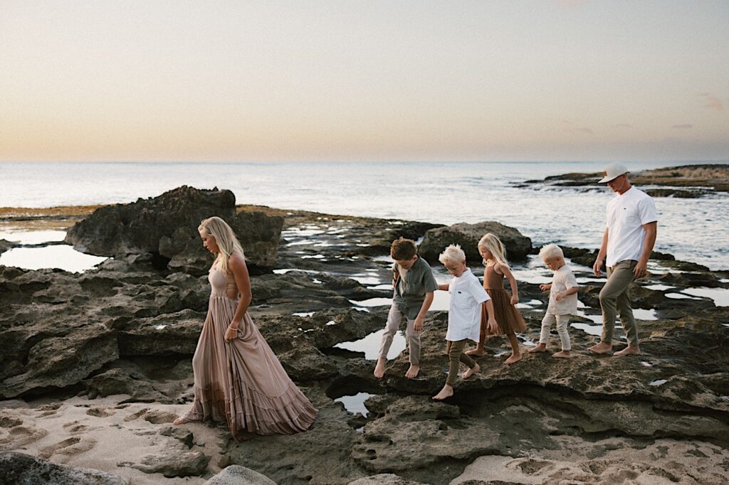 A family of 6 walk along a rocky shore in Hawaii during their family session with the ocean behind them