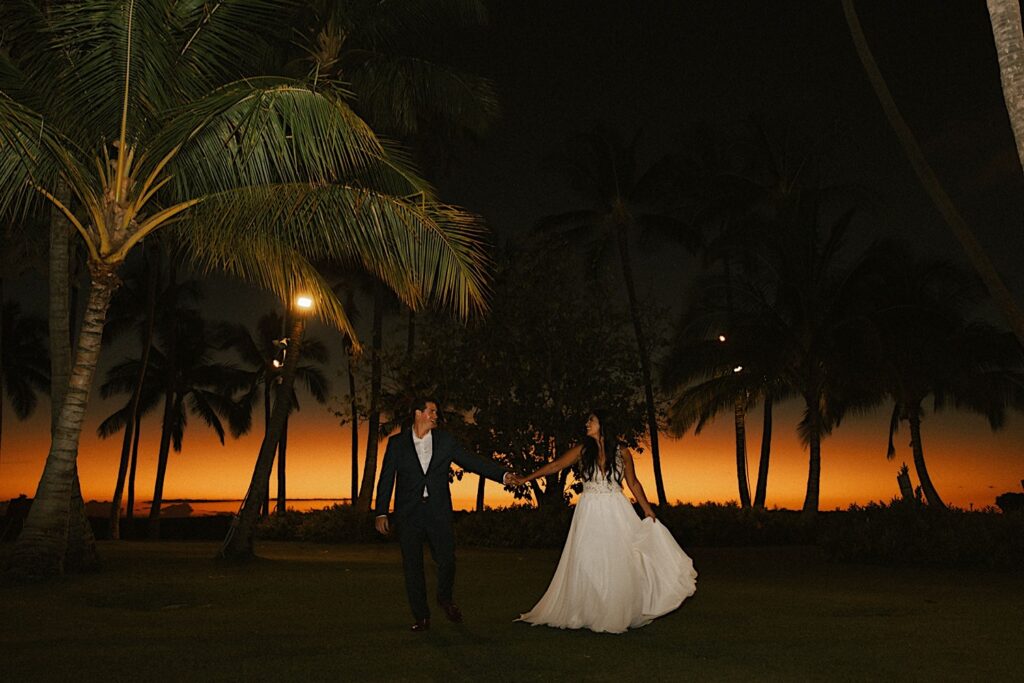 A bride and groom dance in the grass of their wedding venue Lanikuhonua on Oahu surrounded by palm trees as the sun is about set behind them