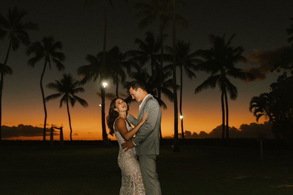 A bride and groom smile and laugh as they dance in the grass of their wedding venue Lanikuhonua on Oahu surrounded by palm trees as the sun is about set behind them