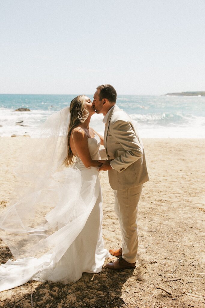 A bride and groom kiss one another in their wedding attire while standing on a beach with the ocean behind them