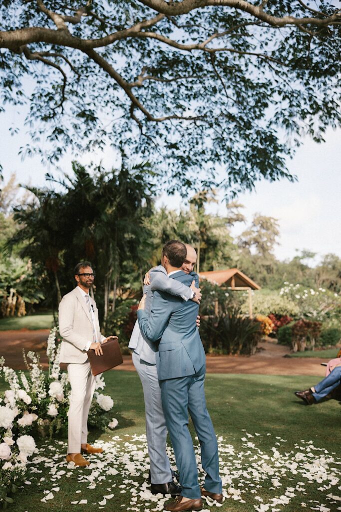 A groom hugs one of his groomsmen at his wedding ceremony space with the officiant standing behind him