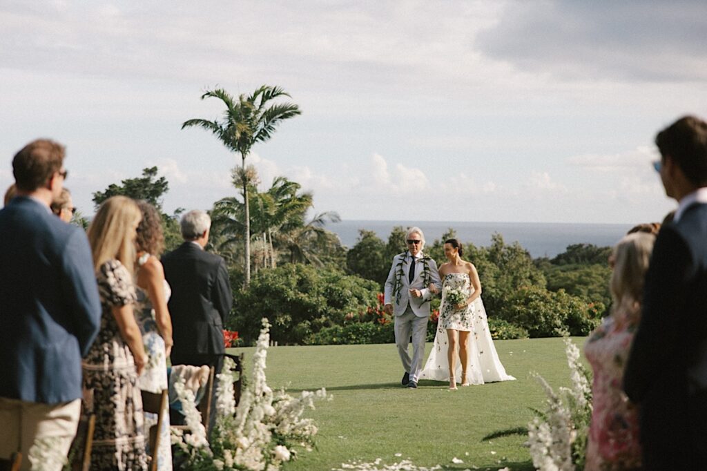 At a wedding ceremony at Kukui'ula on Kauai a bride is walked down the aisle by her father as guests stand and look towards her, behind them is the ocean and palm trees