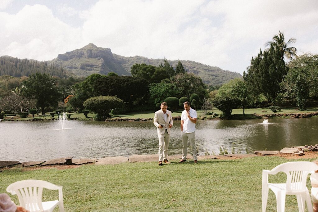 A groom stands with the officiant of his wedding in front of a lake and lush green mountain before an intimate wedding on Kauai