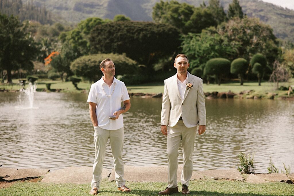 A groom and the officiant of his intimate wedding on Kauai stand in front of a lake as they prepare for the ceremony to begin