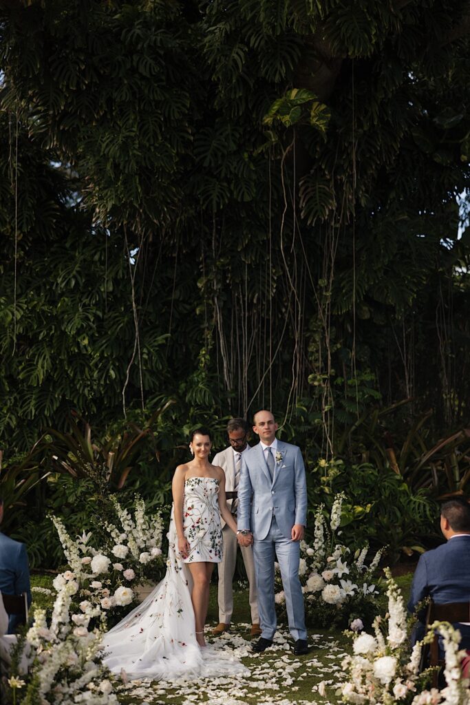 A bride and groom hold hands and face the camera and the guests of their wedding underneath a massive vine covered tree