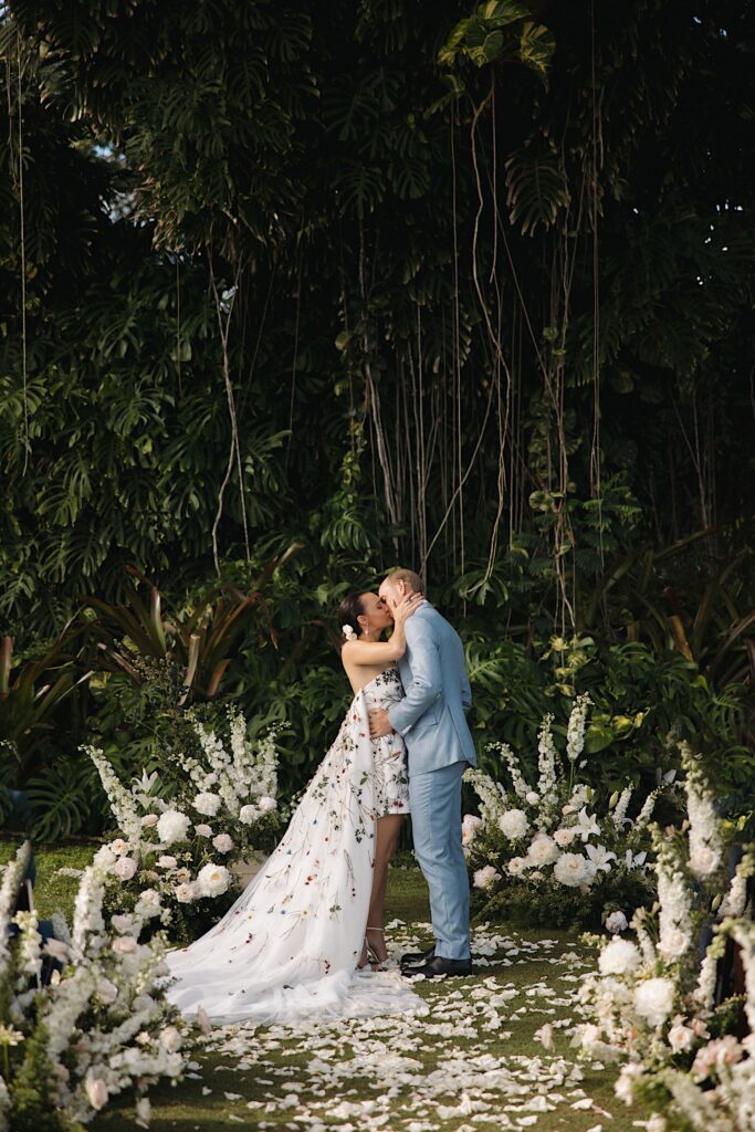A bride and groom kiss one another surrounded by flowers and with a large vine covered tree behind them after their wedding ceremony