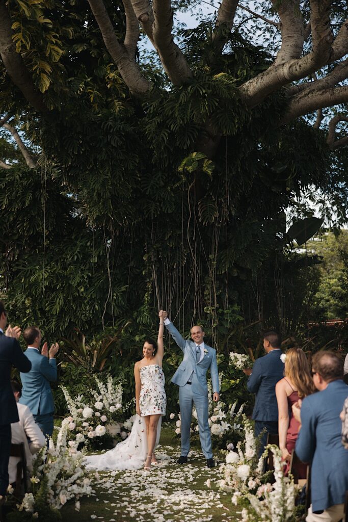 A bride and groom hold hands and raise their hands in the air while smiling at their guests with a massive vine covered tree behind them and white flowers all around them