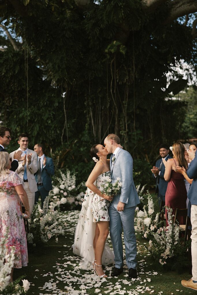 A bride and groom kiss halfway down the aisle after their wedding ceremony with guests on either side of them with a massive vine covered tree behind them