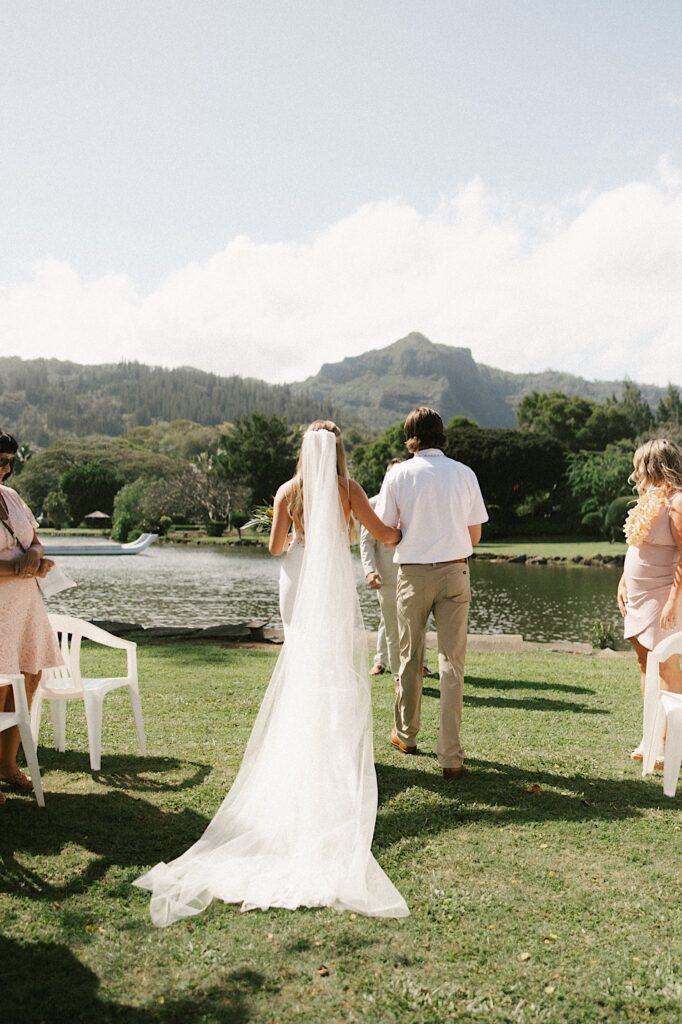 A bride is walked down the aisle away from the camera towards a lake and a mountain in the distance 